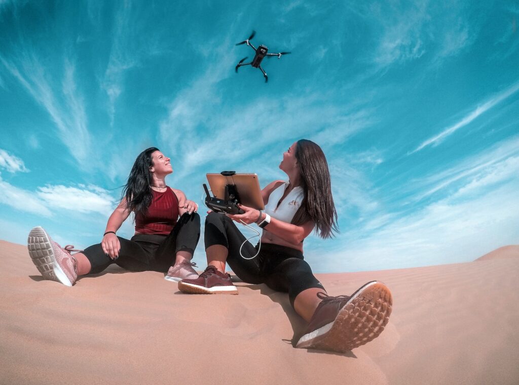 Two Woman Sitting On Sand While Playing Drone
